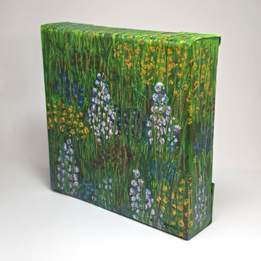 Angled side view of a canvas painting featuring a whimsical scene of bright, happy wildflowers in blue, lavender, red and yellow in a grassy meadow.