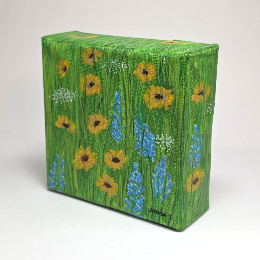 Angled side view of a mini painting of bright little sunflowers with their blue and white wildflower friends in the spring sunshine. A warm grassy meadow adds the perfect background for this joyful mini painting inspired by springtime in central Washington.