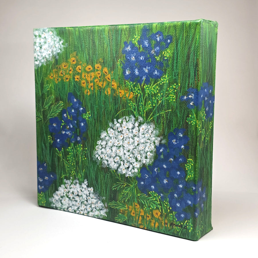 Angled side view of canvas painting of a whimsical scene of peaceful, happy wildflowers in blue, white and yellow against a grassy meadow.