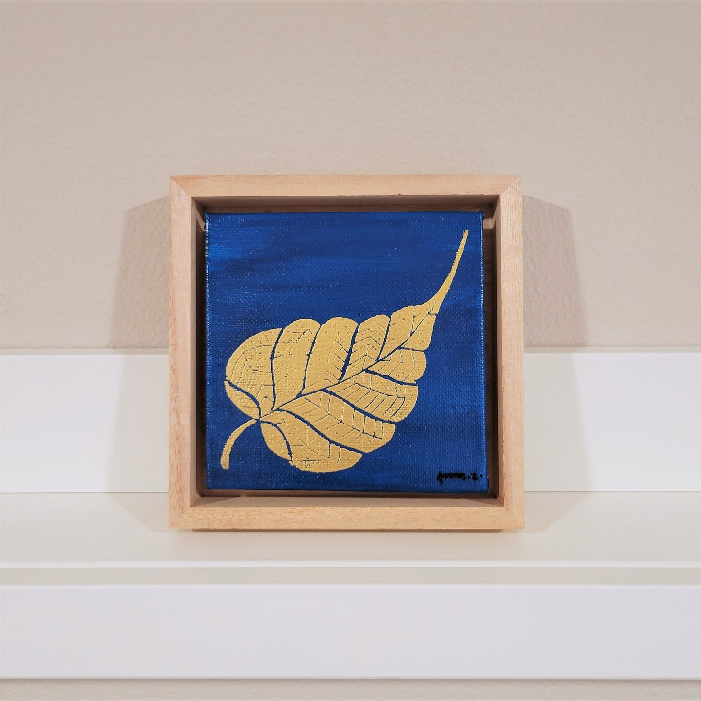 Golden banyan leaf set against a bright blue background on a framed mini-sized canvas panel. Metallic gold colored heat embossing from original carved block design over an acrylic painted background. Mounted in floater frame style within a 5" x 5" rustic wooden box frame with mini sawtooth hanger on reverse. The golden leaf is perfectly complemented by the light colored wooden frame.