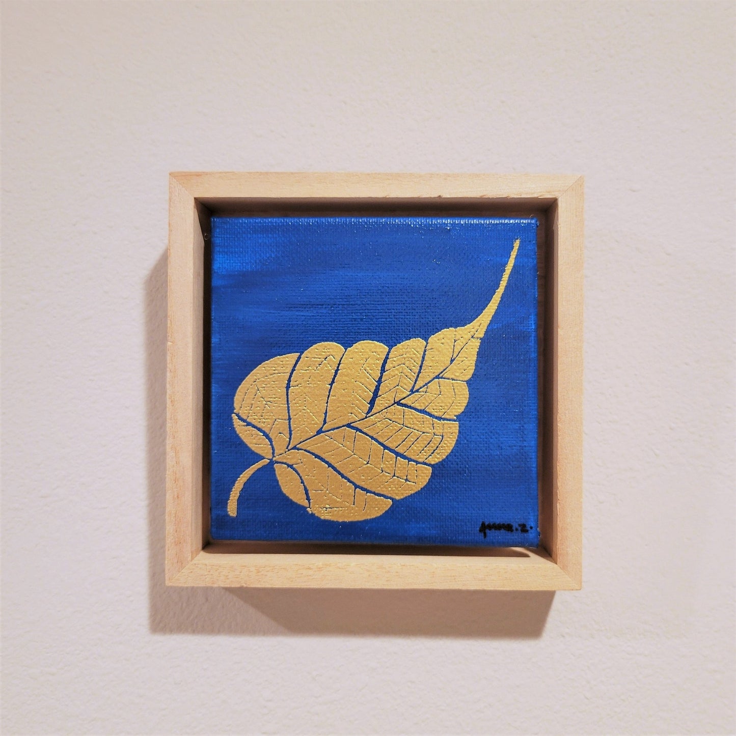 Golden banyan leaf set against a bright blue background on a framed mini-sized canvas panel. Metallic gold colored heat embossing from original carved block design over an acrylic painted background. Mounted in floater frame style within a 5" x 5" rustic wooden box frame.