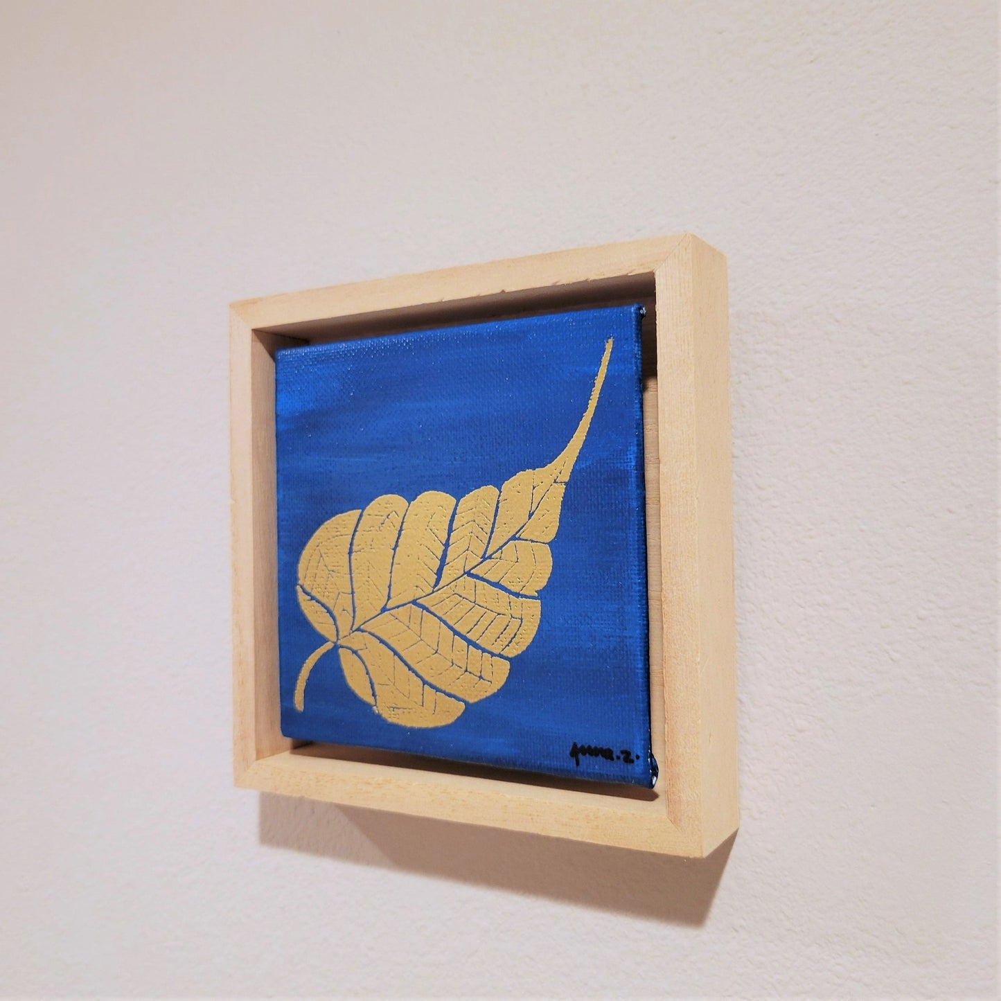 Golden banyan leaf set against a bright blue background on a framed mini-sized canvas panel. Metallic gold colored heat embossing from original carved block design over an acrylic painted background. Mounted in floater frame style within a 5" x 5" rustic wooden box frame with mini sawtooth hanger on reverse. The golden leaf is perfectly complemented by the light colored wooden frame.