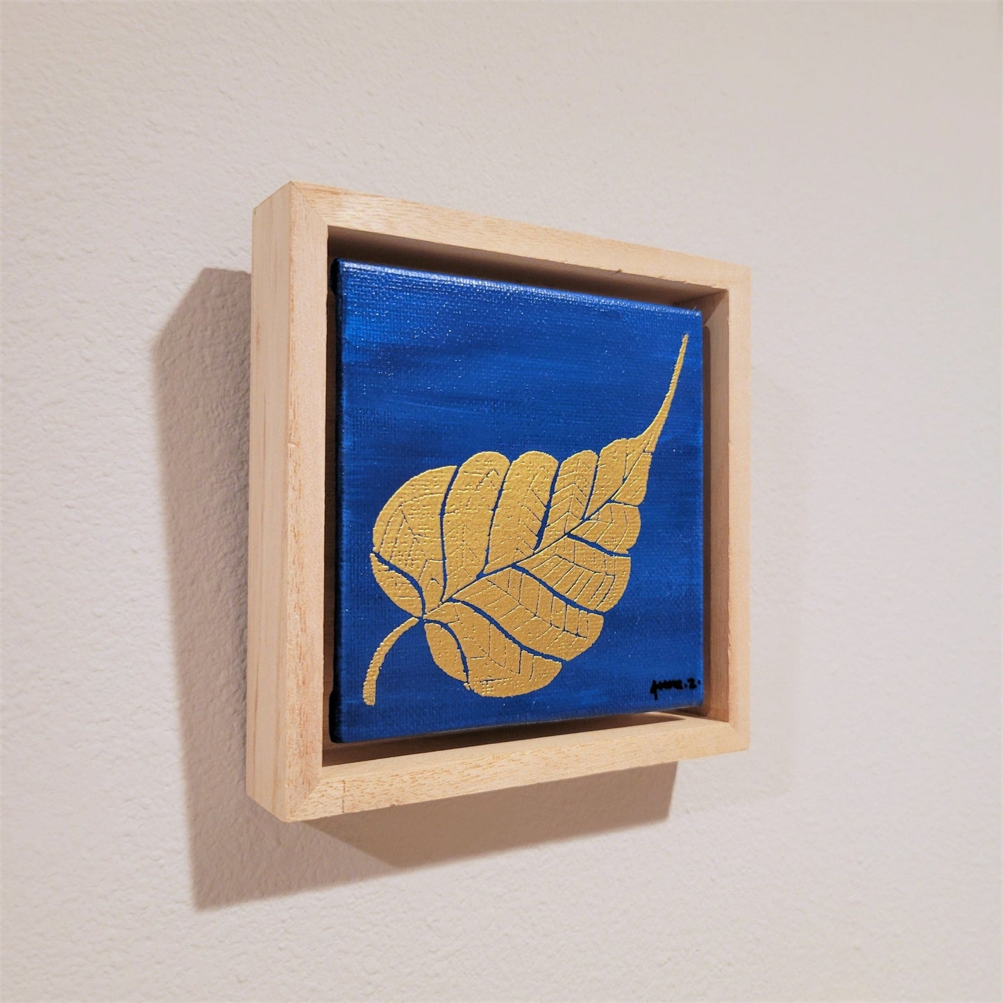 Golden banyan leaf set against a bright blue background on a framed mini-sized canvas panel. Metallic gold colored heat embossing from original carved block design over an acrylic painted background. Mounted in floater frame style within a 5" x 5" rustic wooden box frame with mini sawtooth hanger on reverse. The golden leaf is perfectly complemented by the light colored wooden frame.