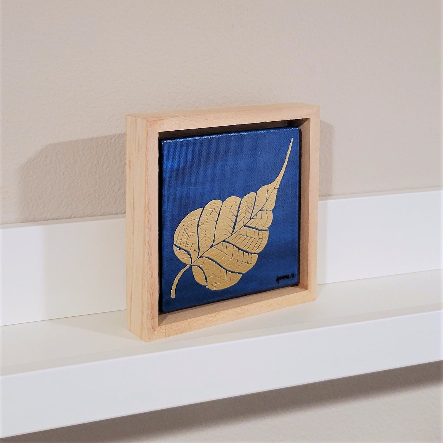 Golden banyan leaf set against a bright blue background on a framed mini-sized canvas panel. Metallic gold colored heat embossing from original carved block design over an acrylic painted background. Mounted in floater frame style within a 5" x 5" rustic wooden box frame with mini sawtooth hanger on reverse. The golden leaf is perfectly complemented by the light colored wooden frame.