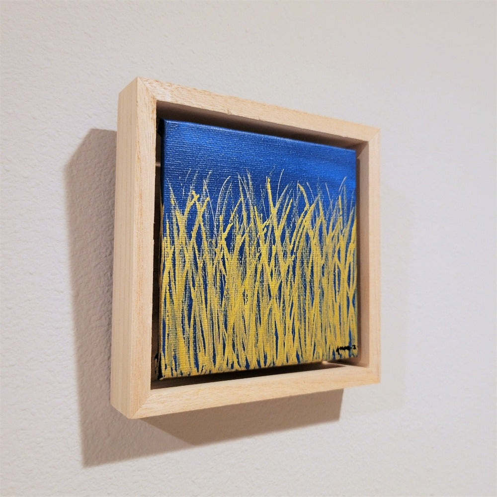 Golden blades of grass set against a bright blue background on a framed mini-sized canvas panel. Metallic gold colored paint over acrylic. Mounted in floater frame style within a 5" x 5" rustic wooden box frame with mini sawtooth hanger on reverse. The golden grass catches daylight beautifully and is perfectly complemented by the light colored wooden frame.