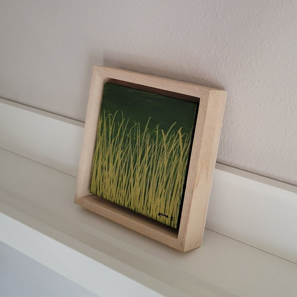 Golden blades of grass set against a deep moss green background on a framed mini-sized canvas panel. Metallic gold colored paint over acrylic. Mounted in floater frame style within a 5" x 5" rustic wooden box frame with mini sawtooth hanger on reverse. The golden grass catches daylight beautifully and is perfectly complemented by the light colored wooden frame.