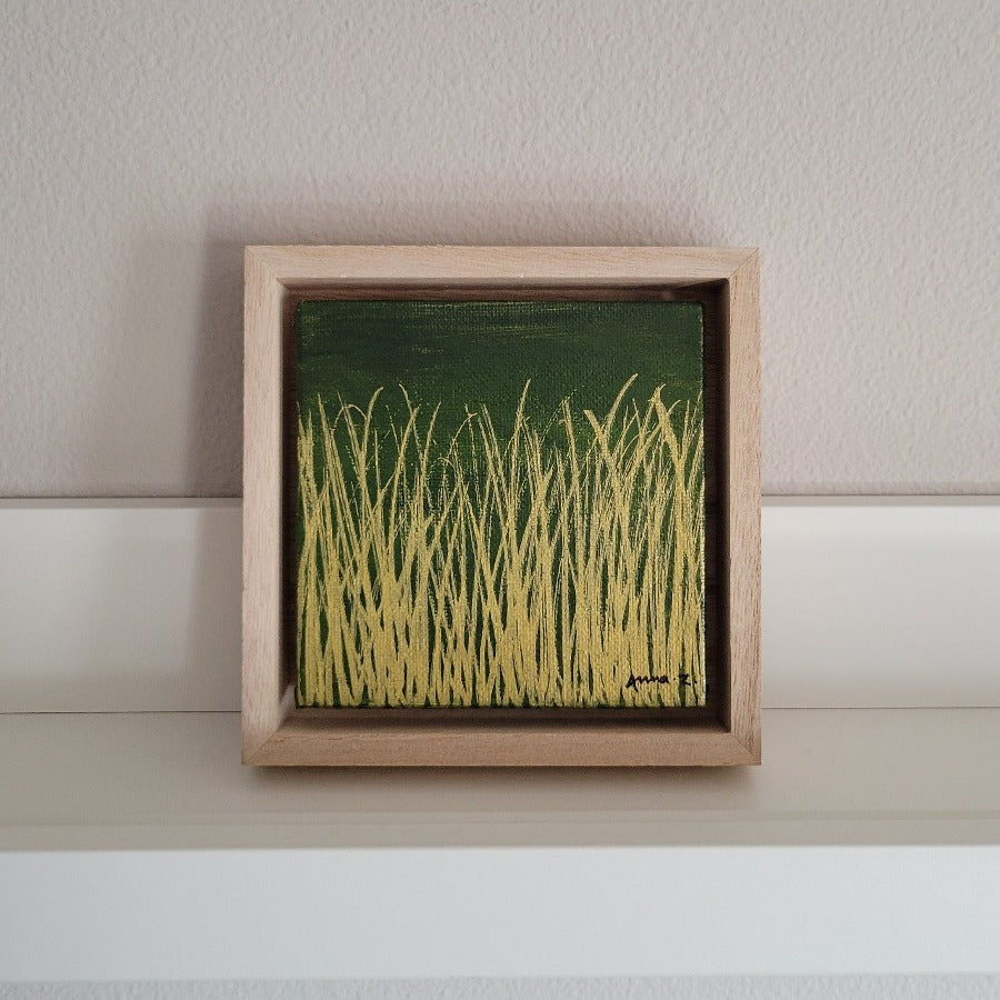 Golden blades of grass set against a deep moss green background on a framed mini-sized canvas panel. Metallic gold colored paint over acrylic. Mounted in floater frame style within a 5" x 5" rustic wooden box frame with mini sawtooth hanger on reverse. The golden grass catches daylight beautifully and is perfectly complemented by the light colored wooden frame.