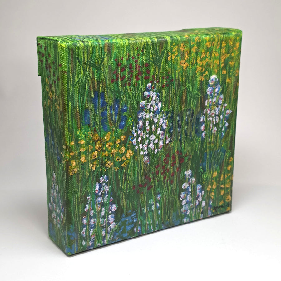 Angled side view of a canvas painting featuring a whimsical scene of bright, happy wildflowers in blue, lavender, red and yellow in a grassy meadow. 