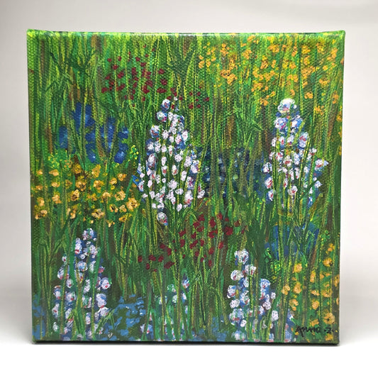 Front view of a canvas painting featuring a whimsical scene of bright, happy wildflowers in blue, lavender, red and yellow in a grassy meadow. 