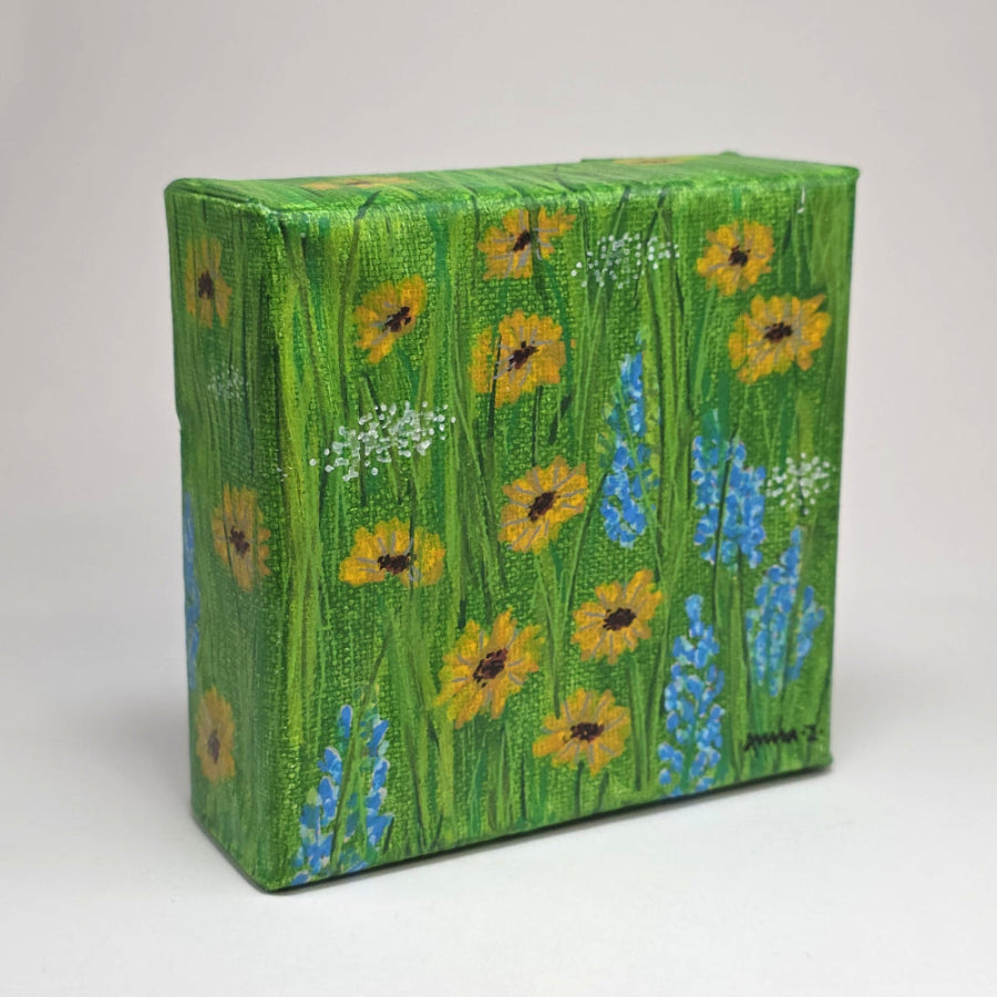 Angled side view of a mini painting of bright little sunflowers with their blue and white wildflower friends in the spring sunshine. A warm grassy meadow adds the perfect background for this joyful mini painting inspired by springtime in central Washington.
