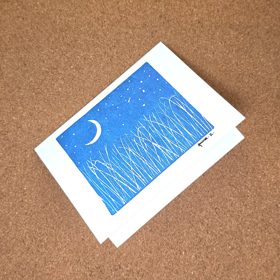 Notecard featuring a digital print reproduction of an original hand-carved block print artwork of a landscape with a crescent moon, starry sky and tall grasses. Blue and white artwork.