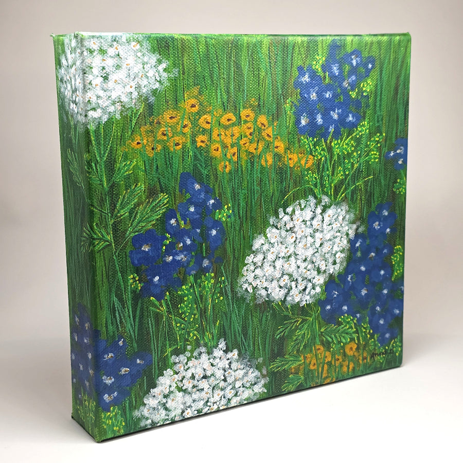 Angled side view of canvas painting of a whimsical scene of peaceful, happy wildflowers in blue, white and yellow against a grassy meadow. 