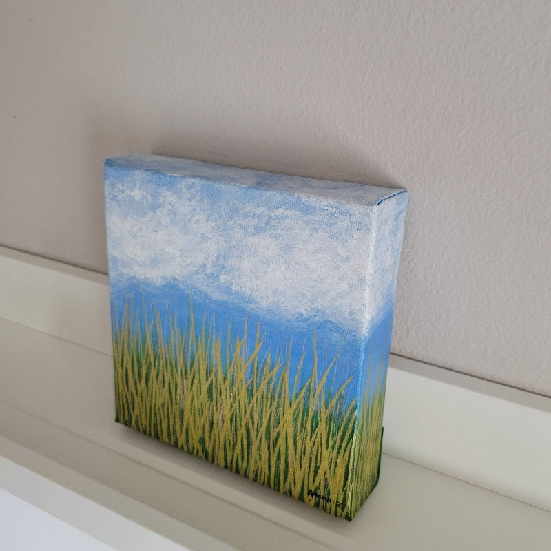 A dreamy mini painting of wispy white clouds against a bright blue sky, with golden grass below and a subtle green background in the distance. The metallic grass shines brilliantly especially in daylight. A cheerful piece capturing nature's beauty in simple details.