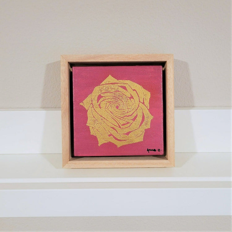 Golden rose set against a bright pink background on a framed mini-sized canvas panel. Metallic gold colored heat embossing from original carved block design, over an acrylic painted background.  Mounted in floater frame style within a 5" x 5" rustic wooden box frame with mini sawtooth hanger on reverse. The golden petals are perfectly complemented by the light colored wooden frame. 