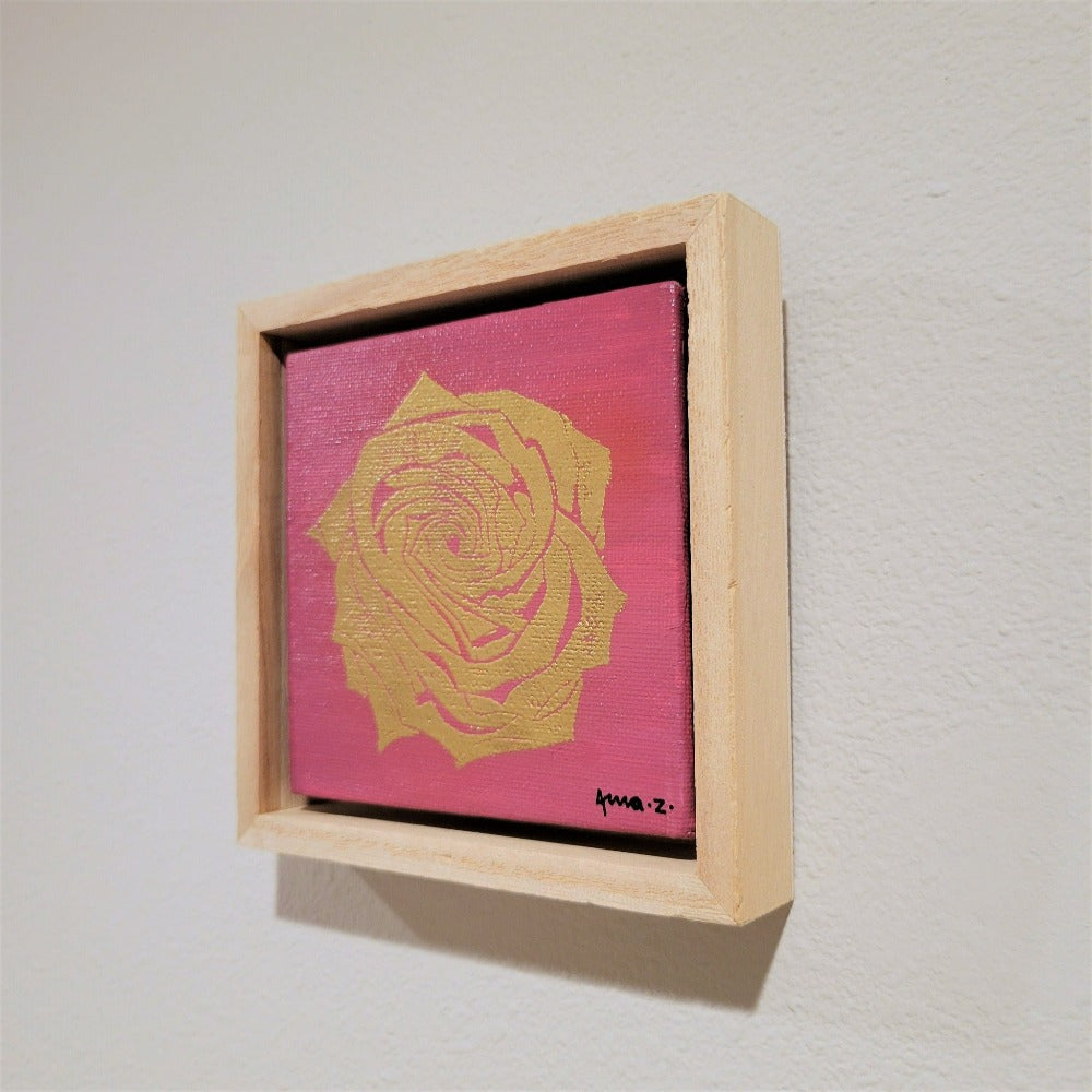 Golden rose set against a bright pink background on a framed mini-sized canvas panel. Metallic gold colored heat embossing from original carved block design, over an acrylic painted background.  Mounted in floater frame style within a 5" x 5" rustic wooden box frame with mini sawtooth hanger on reverse. The golden petals are perfectly complemented by the light colored wooden frame. 