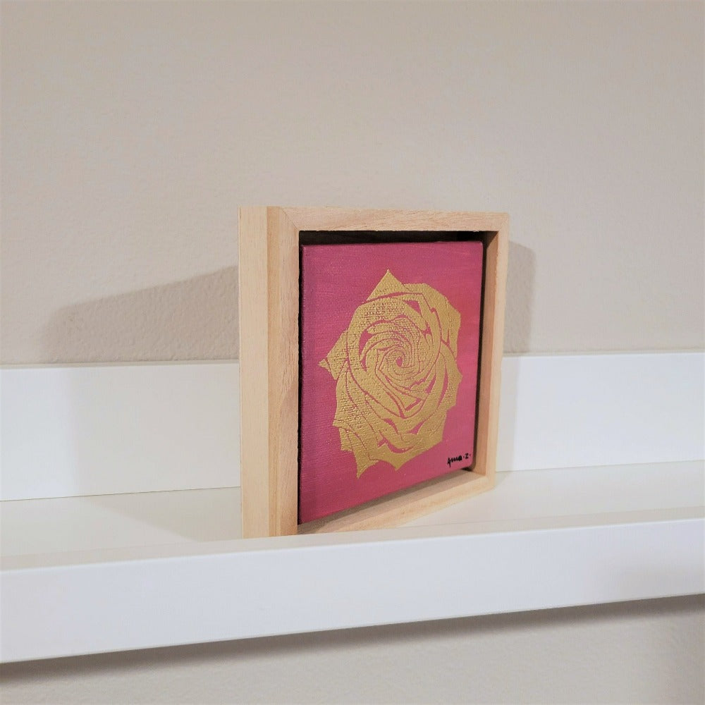 Golden rose set against a bright pink background on a framed mini-sized canvas panel. Metallic gold colored heat embossing from original carved block design, over an acrylic painted background.  Mounted in floater frame style within a 5" x 5" rustic wooden box frame with mini sawtooth hanger on reverse. The golden petals are perfectly complemented by the light colored wooden frame. 