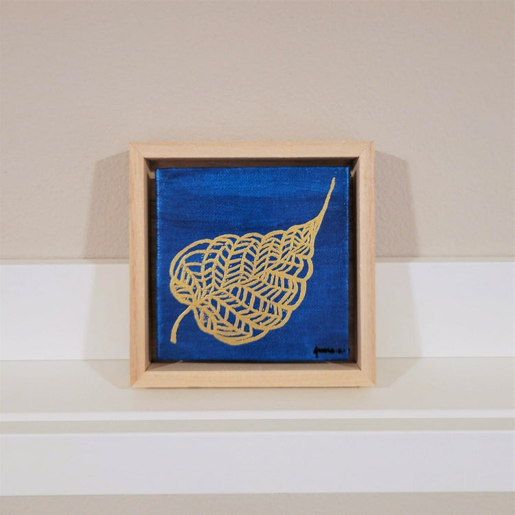 Golden banyan leaf with intricate veins set against a bright blue background on a framed mini-sized canvas panel. Metallic gold colored heat embossing from original carved block design over an acrylic painted background.  Mounted in floater frame style within a 5" x 5" rustic wooden box frame with mini sawtooth hanger on reverse. The golden leaf veins are perfectly complemented by the light colored wooden frame. 