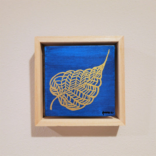 Golden banyan leaf with intricate veins set against a bright blue background on a framed mini-sized canvas panel. Metallic gold colored heat embossing from original carved block design over an acrylic painted background.  Mounted in floater frame style within a 5" x 5" rustic wooden box frame with mini sawtooth hanger on reverse. The golden leaf veins are perfectly complemented by the light colored wooden frame. 