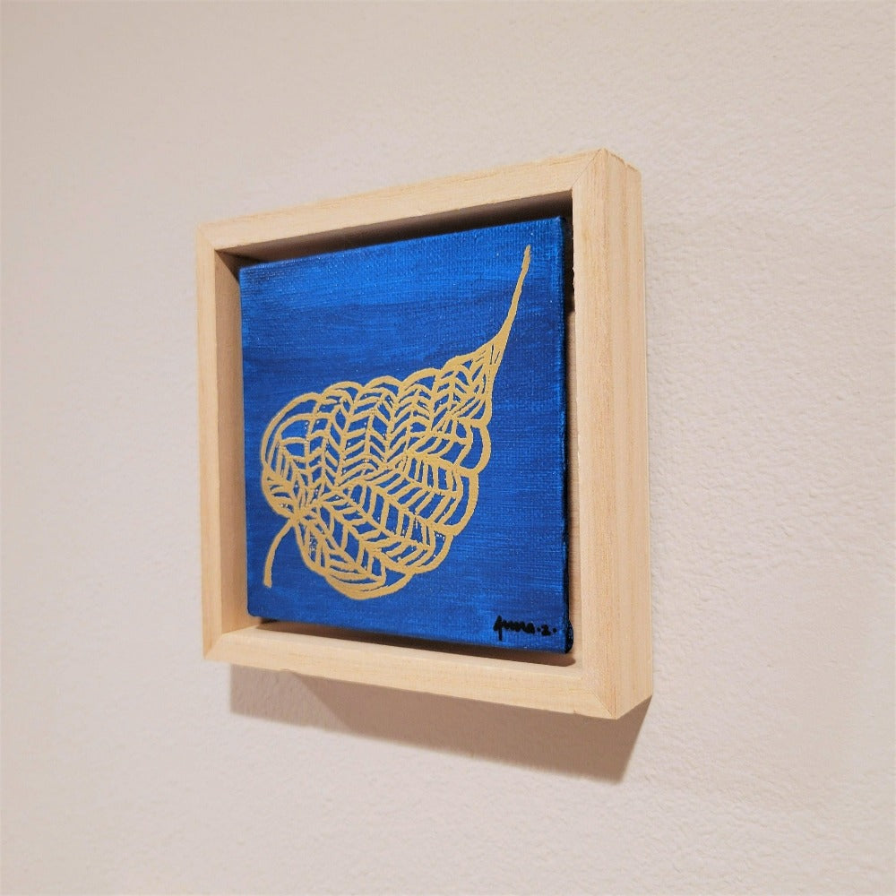 Golden banyan leaf with intricate veins set against a bright blue background on a framed mini-sized canvas panel. Metallic gold colored heat embossing from original carved block design over an acrylic painted background.  Mounted in floater frame style within a 5" x 5" rustic wooden box frame with mini sawtooth hanger on reverse. The golden leaf veins are perfectly complemented by the light colored wooden frame. 