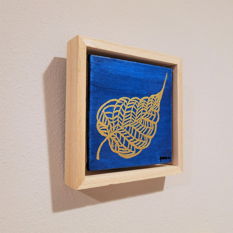 Golden banyan leaf with intricate veins set against a bright blue background on a framed mini-sized canvas panel. Metallic gold colored heat embossing from original carved block design over an acrylic painted background.  Mounted in floater frame style within a 5" x 5" rustic wooden box frame with mini sawtooth hanger on reverse. The golden leaf veins are perfectly complemented by the light colored wooden frame. 