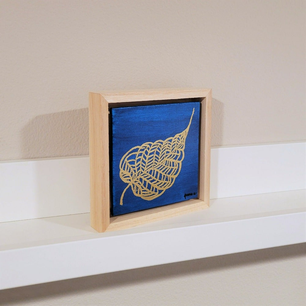 Golden banyan leaf with intricate veins set against a bright blue background on a framed mini-sized canvas panel. Metallic gold colored heat embossing from original carved block design over an acrylic painted background.  Mounted in floater frame style within a 5" x 5" rustic wooden box frame with mini sawtooth hanger on reverse. The golden leaf veins are perfectly complemented by the light colored wooden frame. 