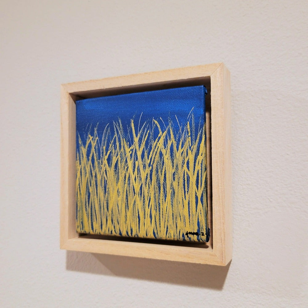 Golden blades of grass set against a bright blue  background on a framed mini-sized canvas panel. Metallic gold colored paint over acrylic.  Mounted in floater frame style within a 5" x 5" rustic wooden box frame with mini sawtooth hanger on reverse. The golden grass catches daylight beautifully and is perfectly complemented by the light colored wooden frame. 
