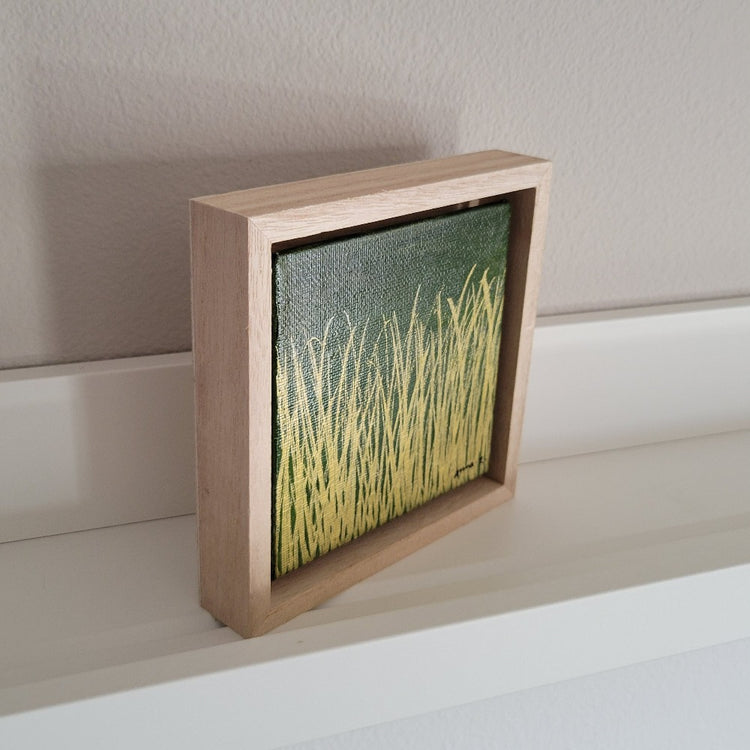 Golden blades of grass set against a deep moss green background on a framed mini-sized canvas panel. Metallic gold colored paint over acrylic. Mounted in floater frame style within a 5" x 5" rustic wooden box frame with mini sawtooth hanger on reverse. The golden grass catches daylight beautifully and is perfectly complemented by the light colored wooden frame. 